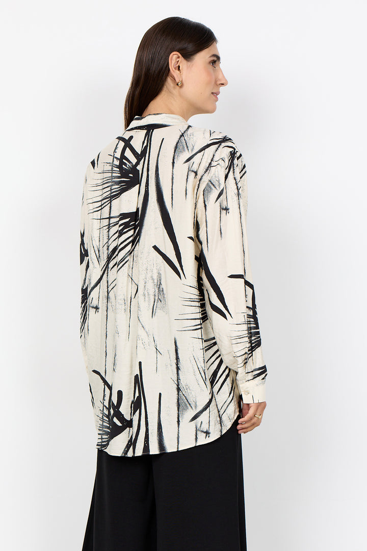 Woman wearing a cream blouse with abstract print and black pants on a white background. 