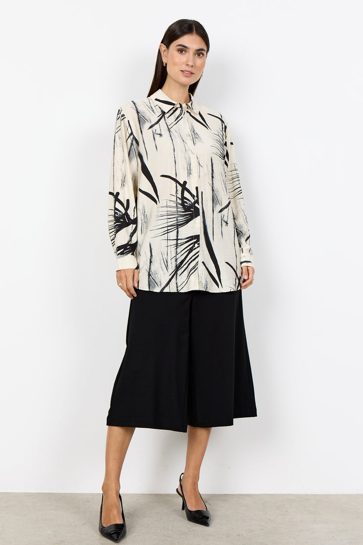 Woman wearing a cream blouse with abstract print and black wide leg pants on a white background. 