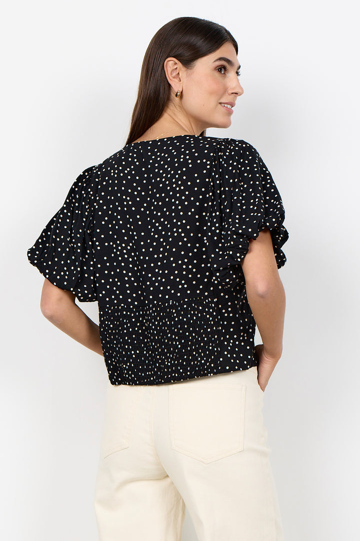 woman wearing a black blouse with white polka dots and balloon sleeves and beige pants on a white background. 
