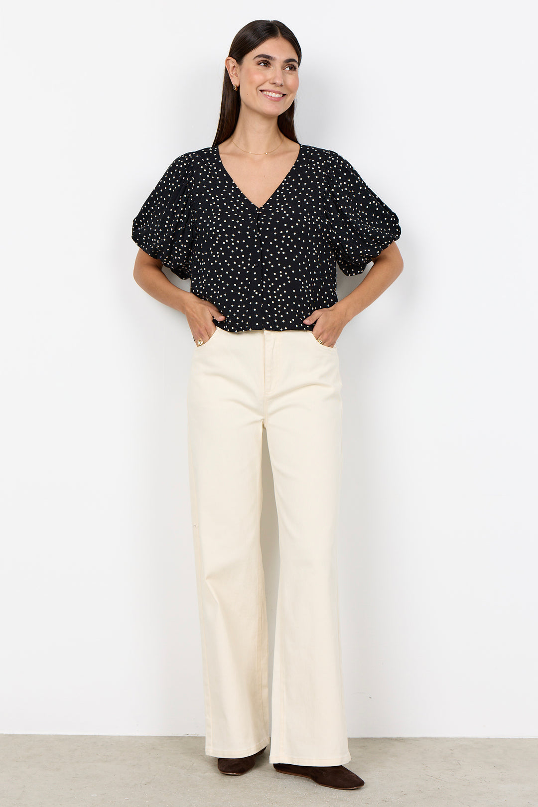 woman wearing a black blouse with white polka dots and balloon sleeves and beige pants on a white background. 
