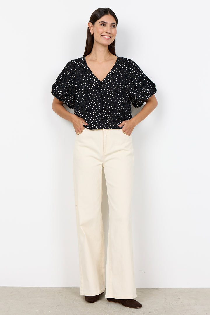 woman wearing a black blouse with white polka dots and balloon sleeves and beige pants on a white background. 