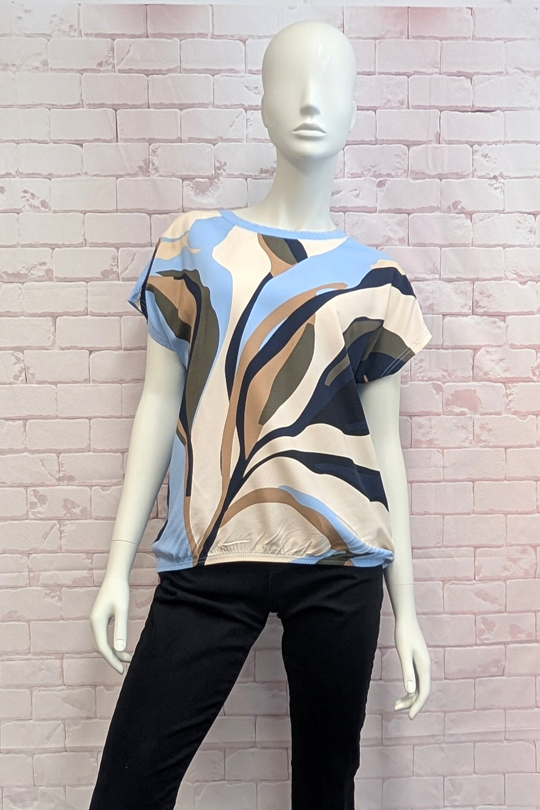 A short-sleeve blouse with a beige, black, brown, and light blue abstract leaf pattern, displayed on a white mannequin against a light pink brick wall.