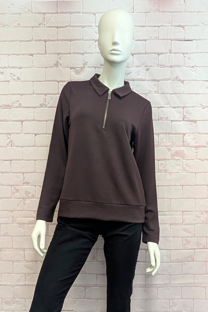A mannequin wearing a dark brown long-sleeve quarter-zip pullover with a polo-style collar, paired with black pants, set against a light brick wall background.
