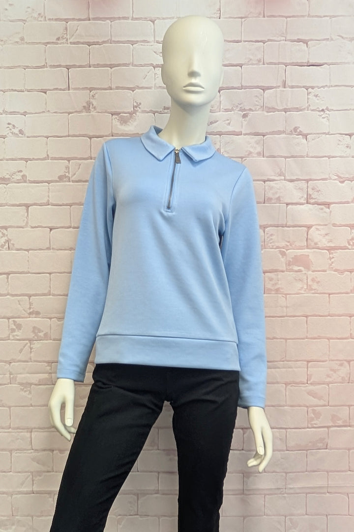 A light blue long-sleeve polo-style top displayed on a mannequin, paired with black pants, set against a light pink brick wall background.