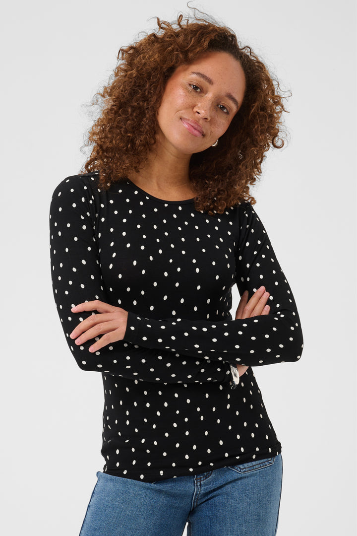 A woman with curly brown hair wearing a black long-sleeve top with white polka dots and blue jeans, posing against a light gray studio background.