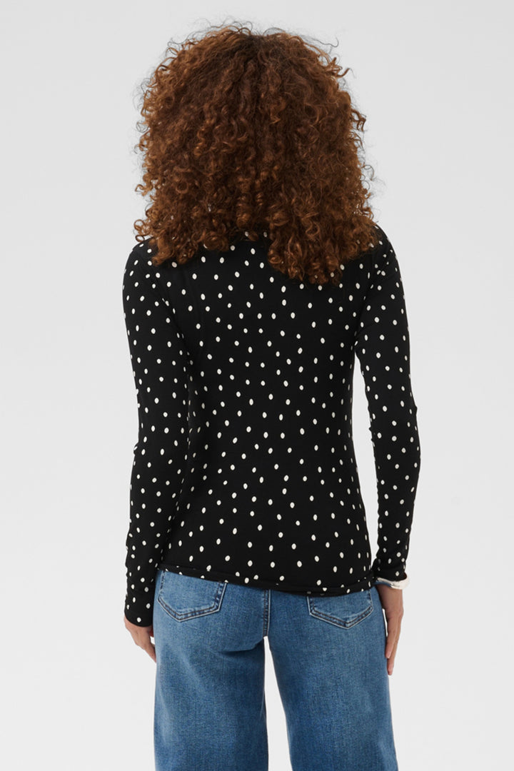 A woman viewed from the back wearing a black long-sleeve top with a white polka dot pattern and blue jeans, set against a plain light gray studio background.