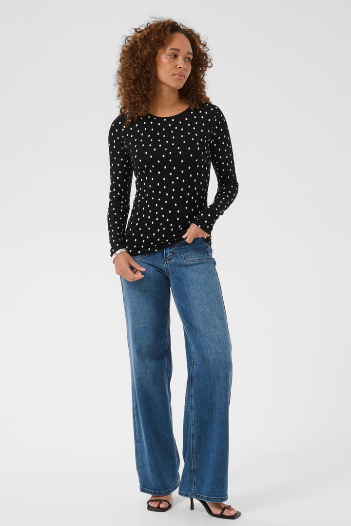 A woman modeling a black long-sleeve top with a white polka-dot pattern, paired with blue wide-leg jeans and black sandals, against a light gray studio background.