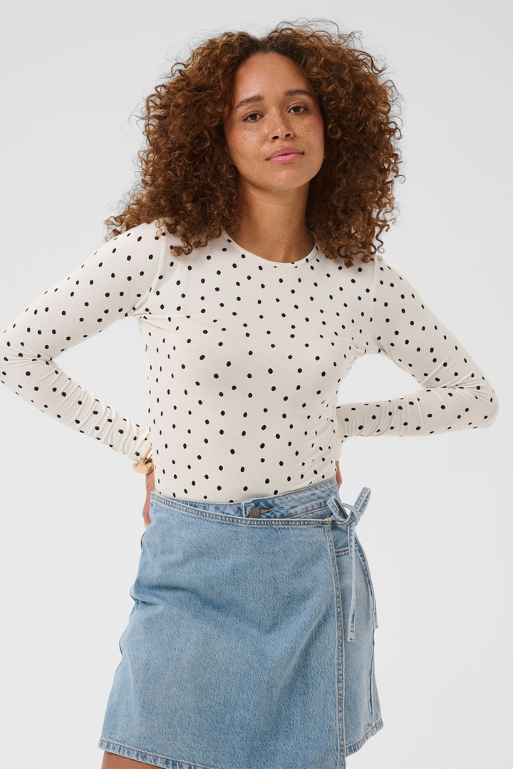 A woman with curly brown hair wearing a white long-sleeve top with black polka dots and a light blue denim mini skirt, posing against a light gray studio background.
