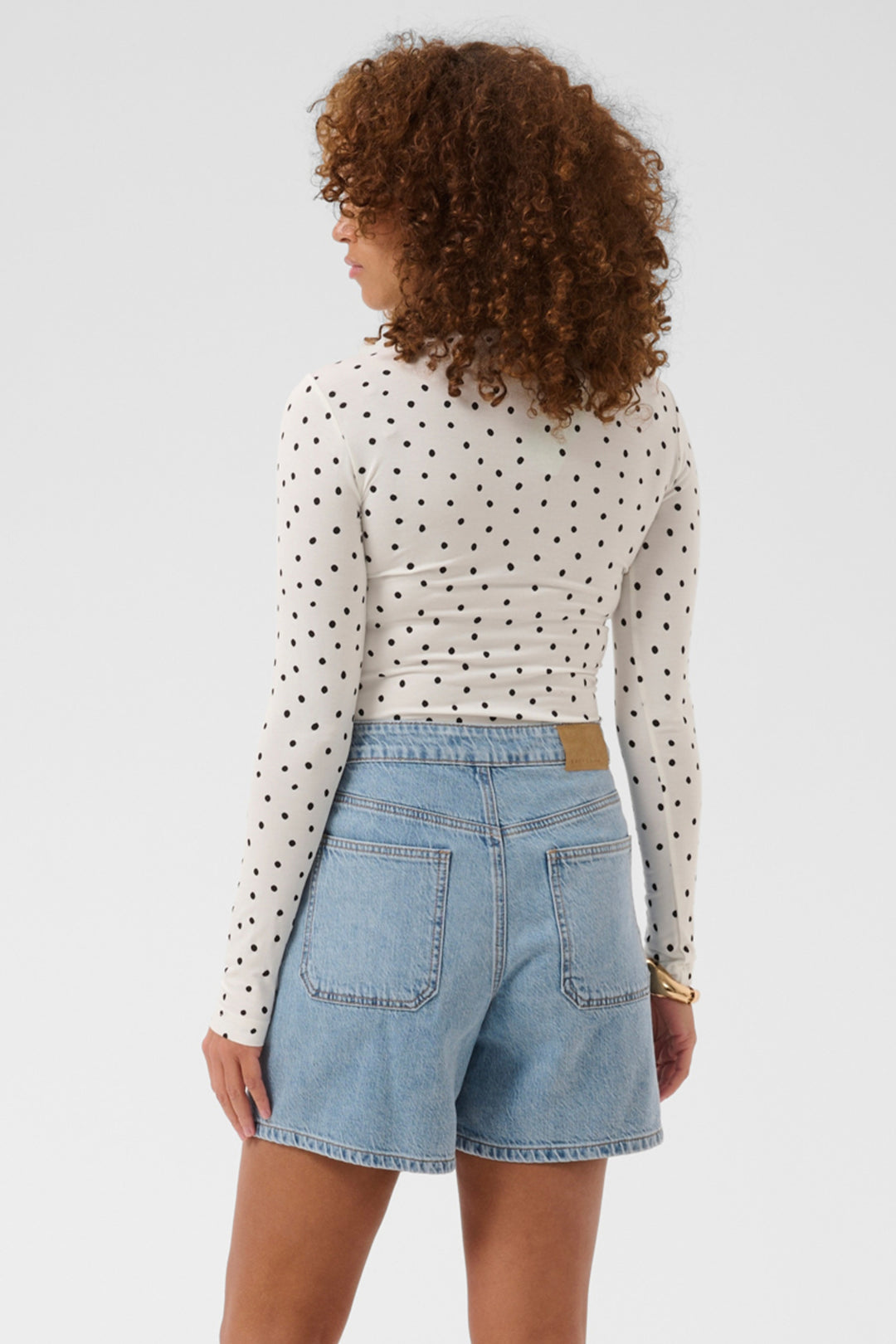 A woman shown from the back wearing a white long-sleeve crop top with black polka dots and light blue denim shorts against a light gray studio background.