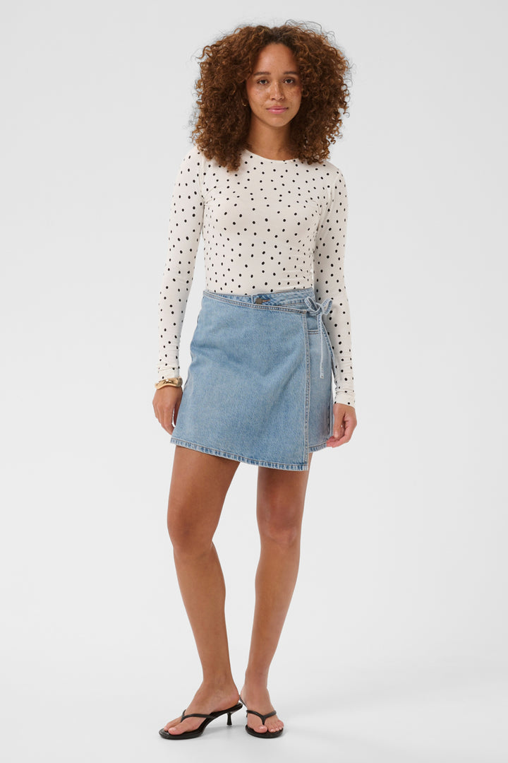 A woman modeling a white long-sleeve knit top with black polka dots and a light blue denim mini skirt, paired with black heeled sandals, against a plain light gray studio background.