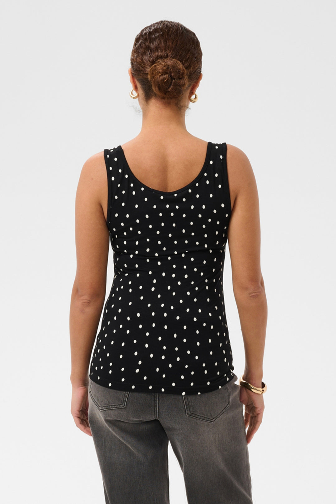 A woman viewed from the back wearing a black sleeveless tank top with a white polka-dot pattern and gray jeans, against a light gray studio background.