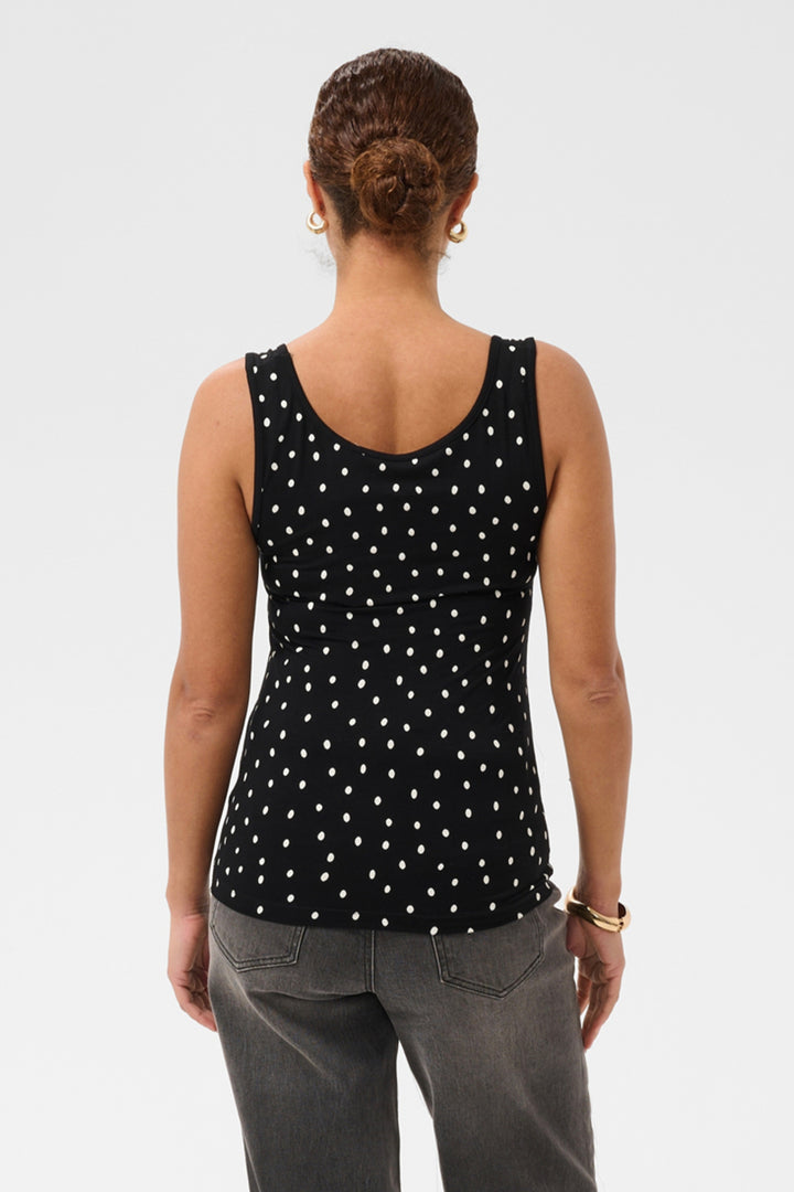 A woman viewed from the back wearing a black sleeveless tank top with a white polka-dot pattern and gray jeans, against a light gray studio background.