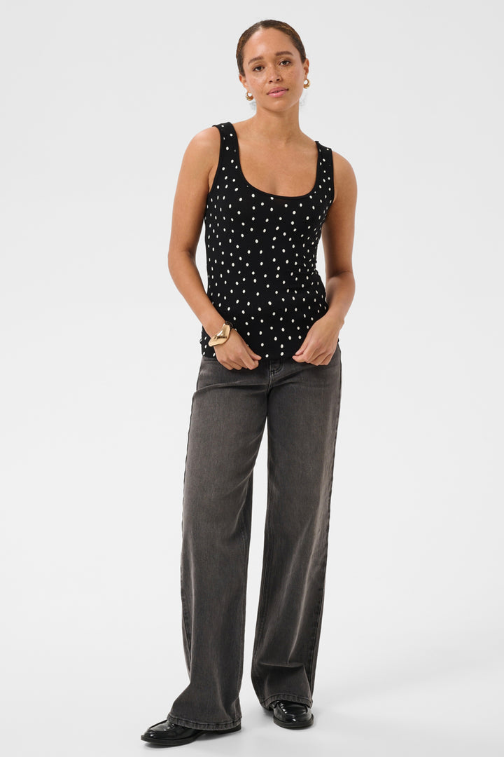A woman modeling a black sleeveless tank top with white polka dots and dark gray wide-leg jeans against a plain light gray studio background.