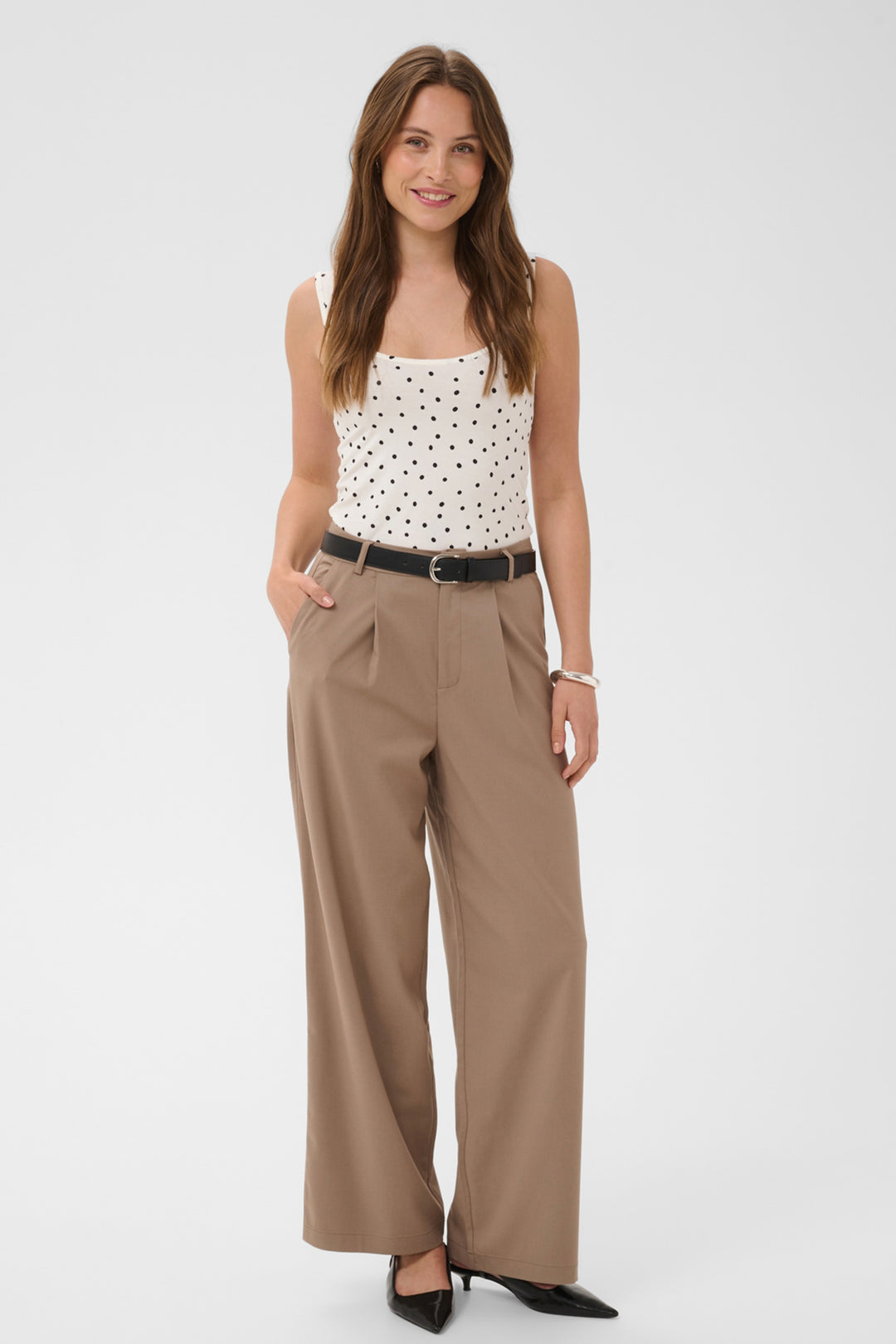 A woman modeling a white sleeveless top with black polka dots, paired with high-waisted, wide-leg taupe pants, a black belt, and black pointed-toe heels, against a light gray studio background.