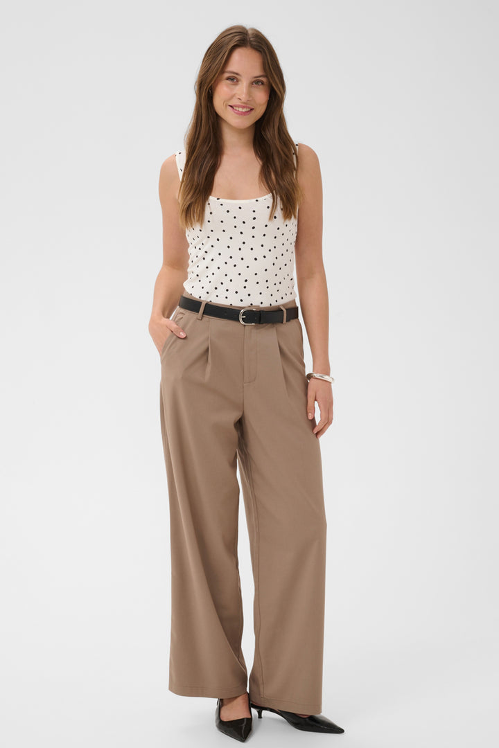 A woman modeling a white sleeveless top with black polka dots, paired with high-waisted, wide-leg taupe pants, a black belt, and black pointed-toe heels, against a light gray studio background.