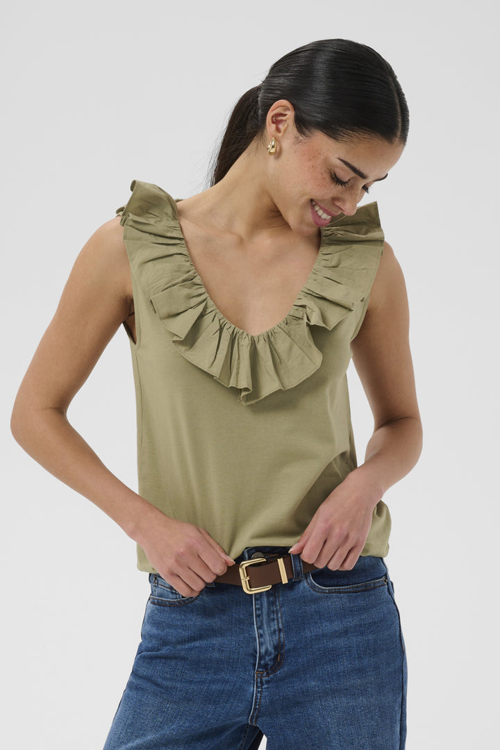A woman wearing a sleeveless olive green ruffled top and blue jeans, looking down and smiling. The photo is taken against a plain light gray studio background.