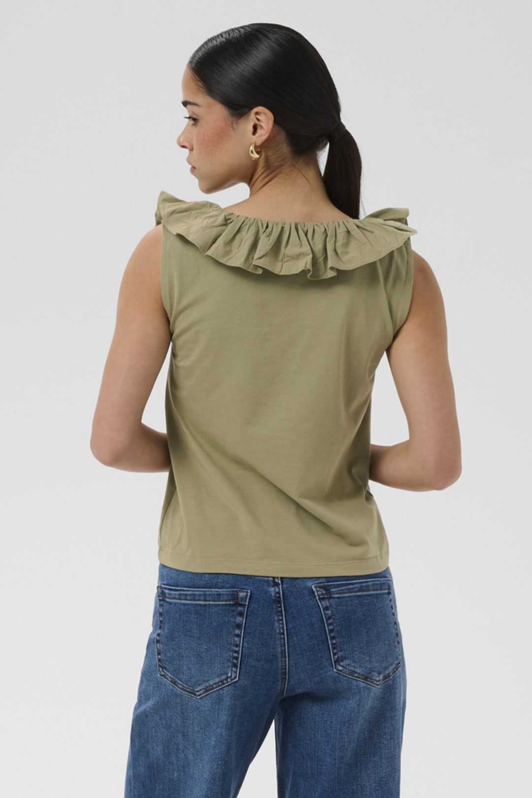 A woman shown from the back wearing an olive green sleeveless top with a ruffled collar and blue jeans, against a light gray studio background.