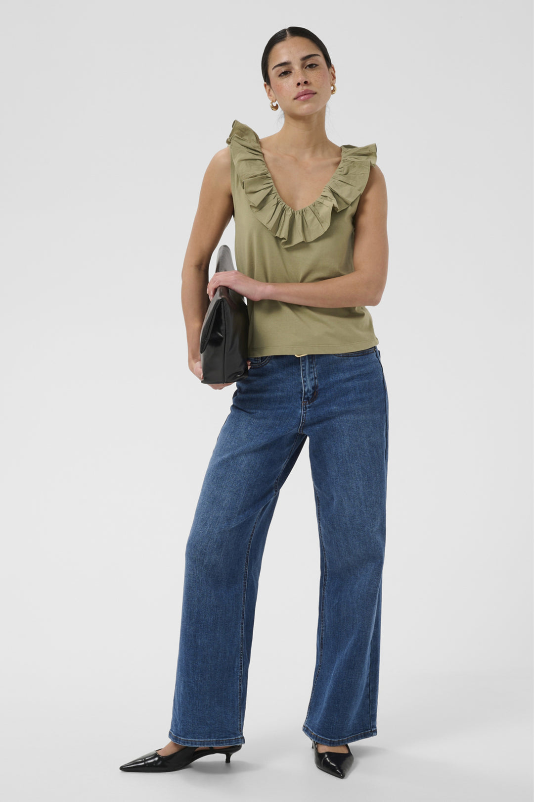 A model stands against a light gray studio background wearing an olive green ruffled sleeveless top, wide-leg blue jeans, and black pointed flats, while holding a black clutch.