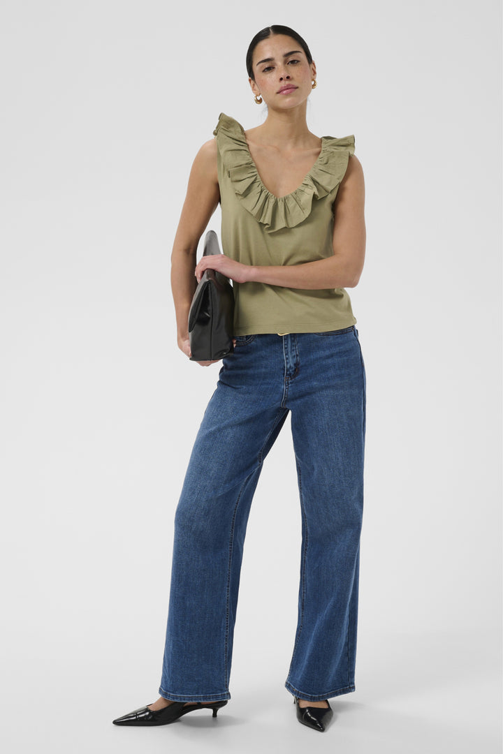 A model stands against a light gray studio background wearing an olive green ruffled sleeveless top, wide-leg blue jeans, and black pointed flats, while holding a black clutch.