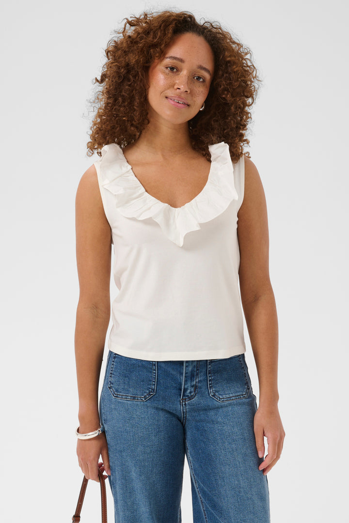 A woman with curly brown hair wearing a white sleeveless top with a ruffled v-neck collar and blue jeans, standing against a light gray studio background.