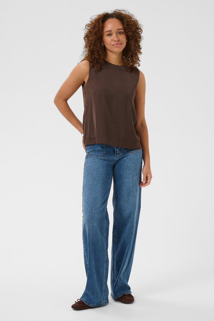 A woman with curly brown hair stands against a light gray studio background wearing a sleeveless dark brown top and wide-leg blue jeans.