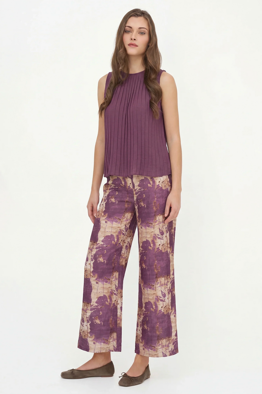 A woman modeling a sleeveless pleated top in dusty purple paired with wide-leg trousers featuring a purple and beige abstract print, against a plain white studio background.