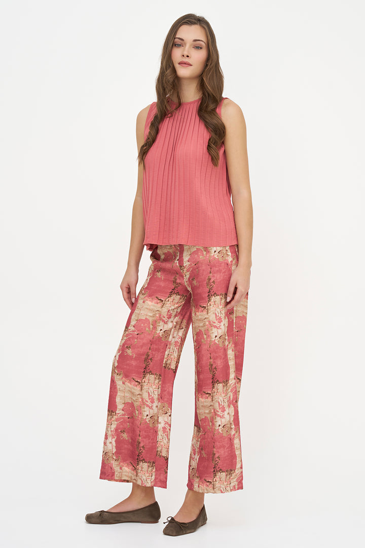 A woman modeling a sleeveless pleated pink top and wide-leg trousers with a pink and beige abstract print, paired with dark brown flats, against a plain white studio background.