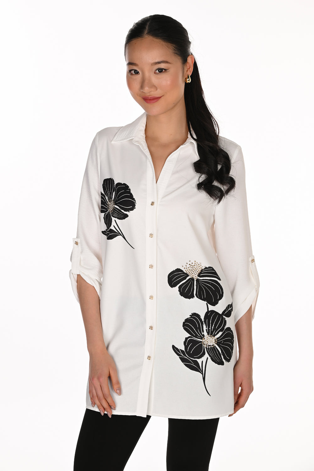 A woman modeling a white button-up shirt with black floral embroidery and small rhinestone accents, paired with black pants, against a plain white background.
