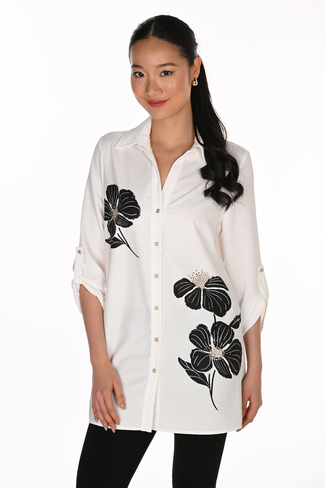 A woman modeling a white button-up shirt with black floral embroidery and small rhinestone accents, paired with black pants, against a plain white background.