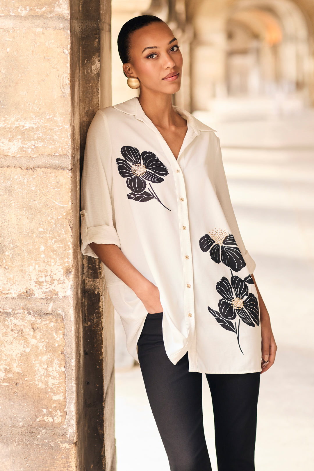 A woman leans against a stone pillar in a sunlit corridor, wearing a loose white button-front shirt with large black floral embroidery and black pants.