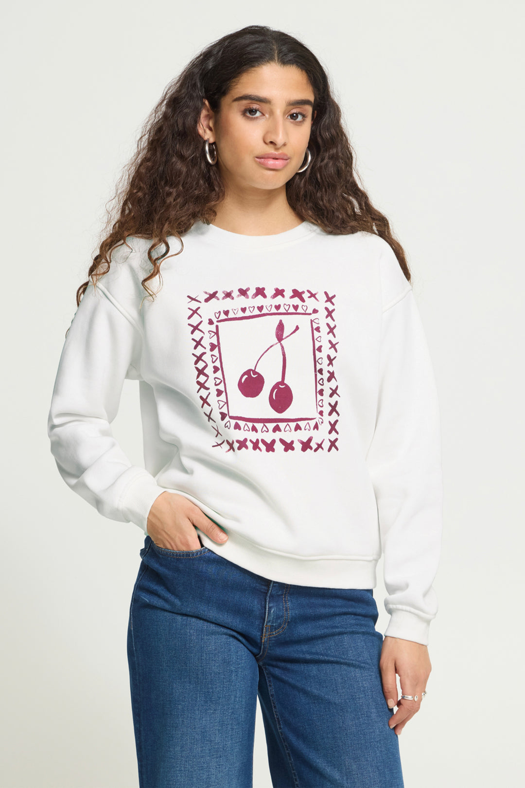 A woman wearing a white crewneck sweatshirt with a maroon graphic of cherries and the word &