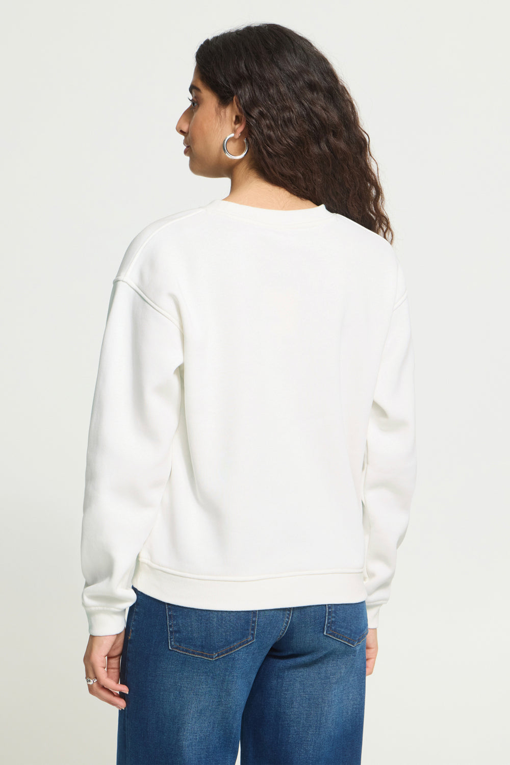 A woman shown from the back wearing a white crewneck sweatshirt and blue jeans against a plain light background.