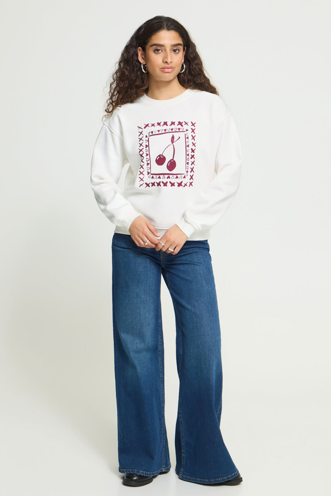 A model wearing a white crewneck sweatshirt with a red cherry graphic print, paired with blue wide-leg jeans, against a plain light gray studio background.