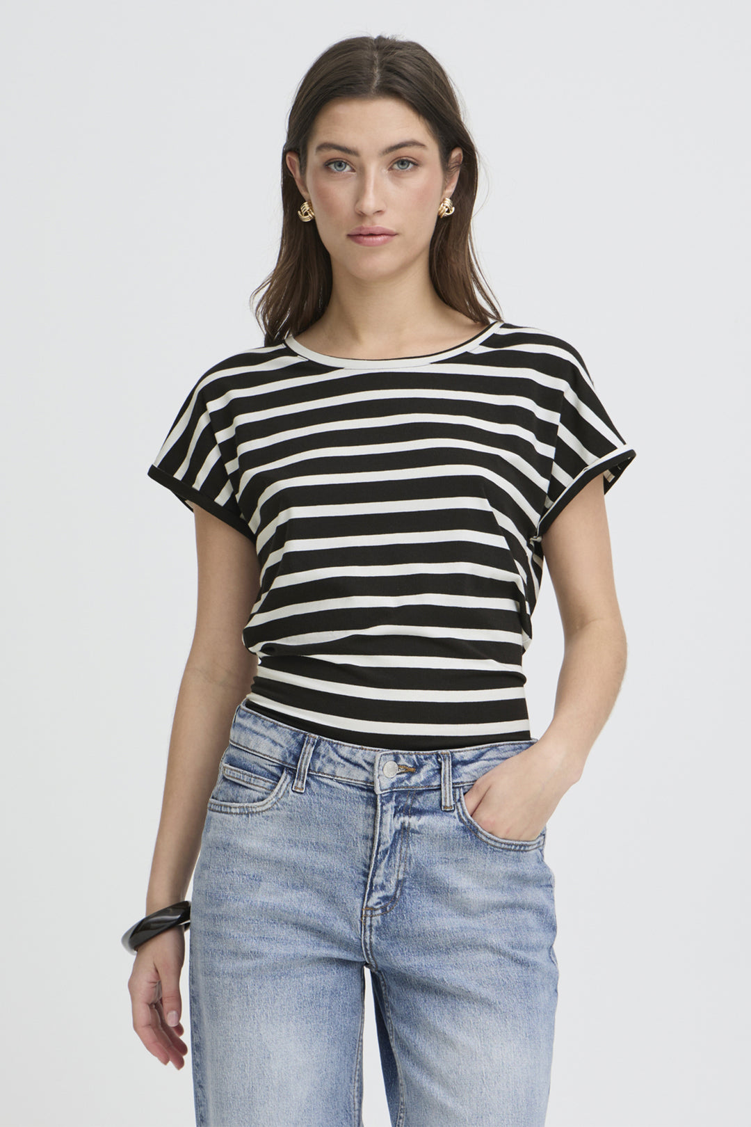 A woman modeling a black and white horizontally striped short-sleeve crop top with light blue jeans, set against a plain light gray studio background.