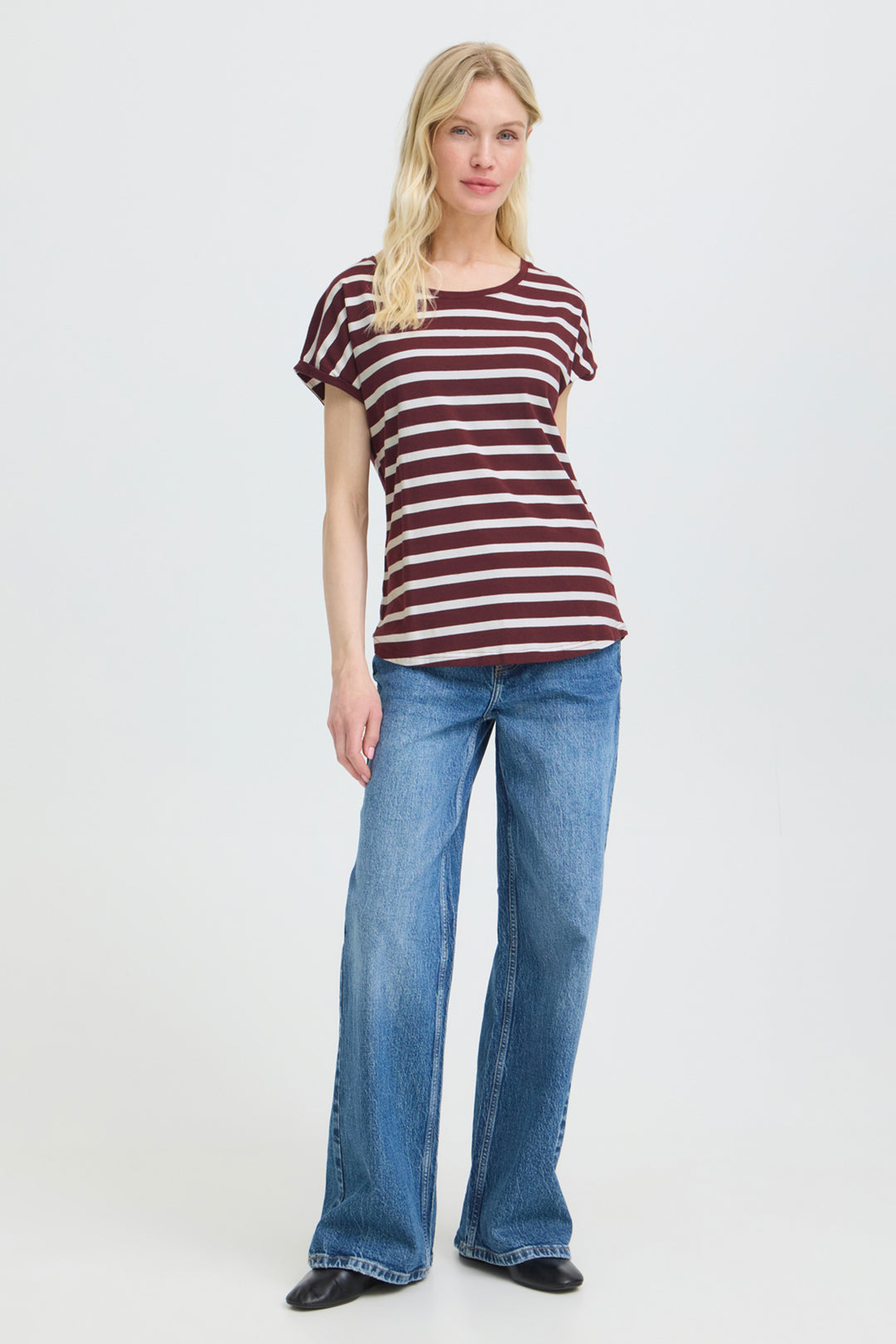 A woman modeling a short-sleeve crewneck t-shirt with horizontal burgundy and white stripes, paired with wide-leg blue jeans and black shoes, against a plain light gray studio background.