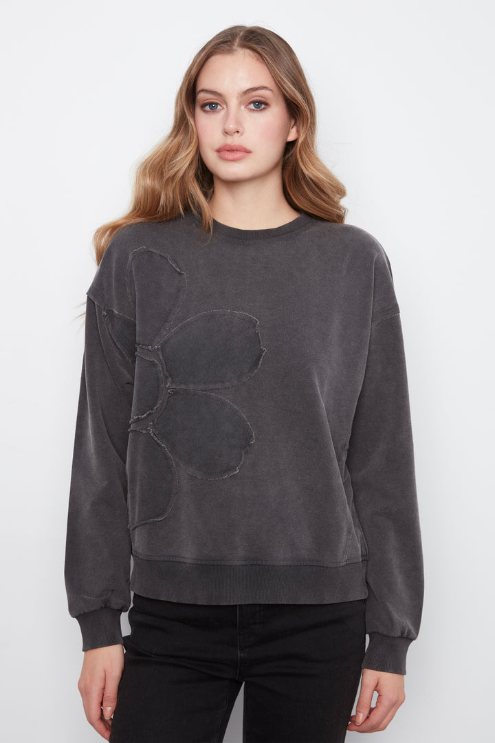 A woman modeling a dark gray washed crewneck sweatshirt with a large tonal abstract patch on the front, paired with black pants, against a plain light gray background.