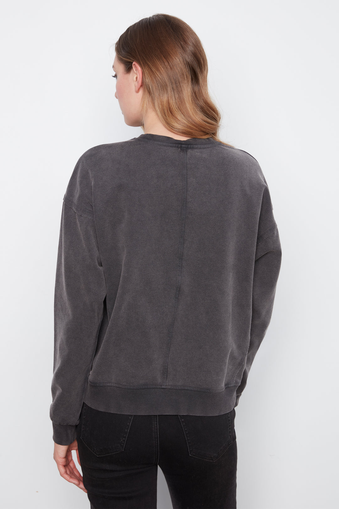 A woman shown from the back wearing a dark gray washed crewneck sweatshirt and black pants, photographed against a plain white studio background.