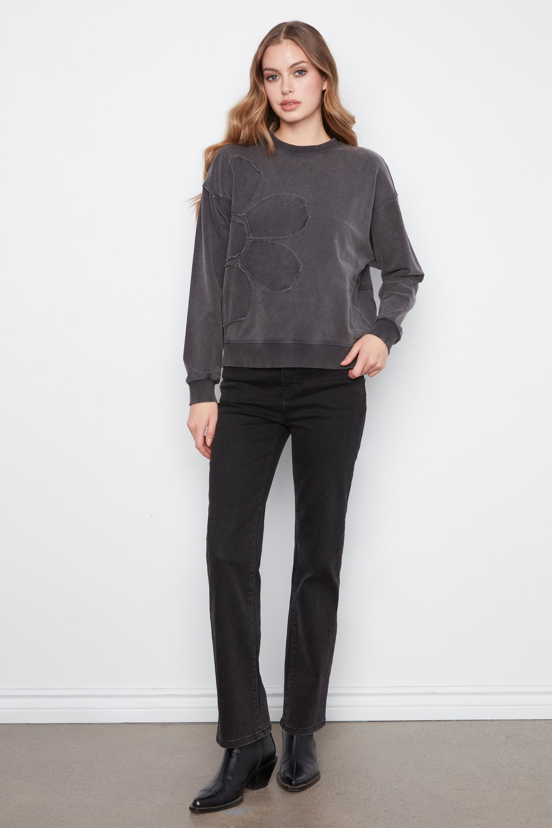 A woman modeling a dark gray crewneck sweatshirt with a tonal geometric pattern on the front, paired with black jeans and black leather ankle boots, standing against a white wall.