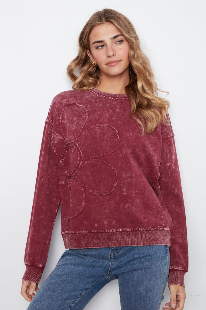 A woman wearing a washed, dusty rose long-sleeve sweatshirt with a large tonal floral applique on the front, paired with blue jeans, against a plain white studio background.
