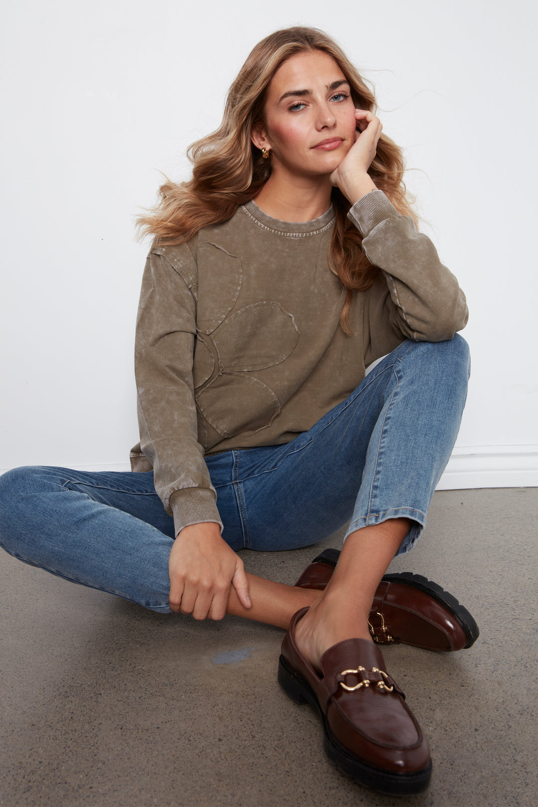 A woman sits cross-legged on a gray concrete floor against a white wall, wearing a taupe-brown textured crewneck sweatshirt, blue jeans, and brown leather loafers with gold hardware.