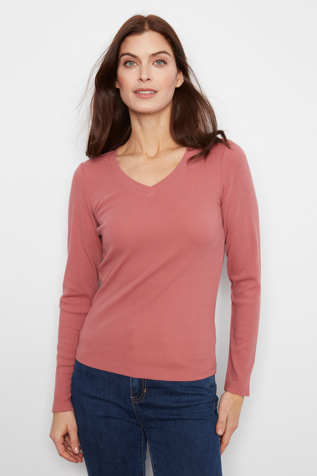 A woman wearing a dusty rose long-sleeve V-neck knit top and dark blue jeans, standing against a plain light gray studio background.