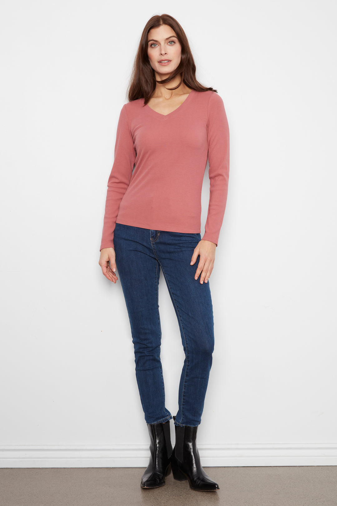 A woman modeling a dusty rose long-sleeve V-neck knit top with dark blue skinny jeans and black ankle boots, standing against a plain white wall.