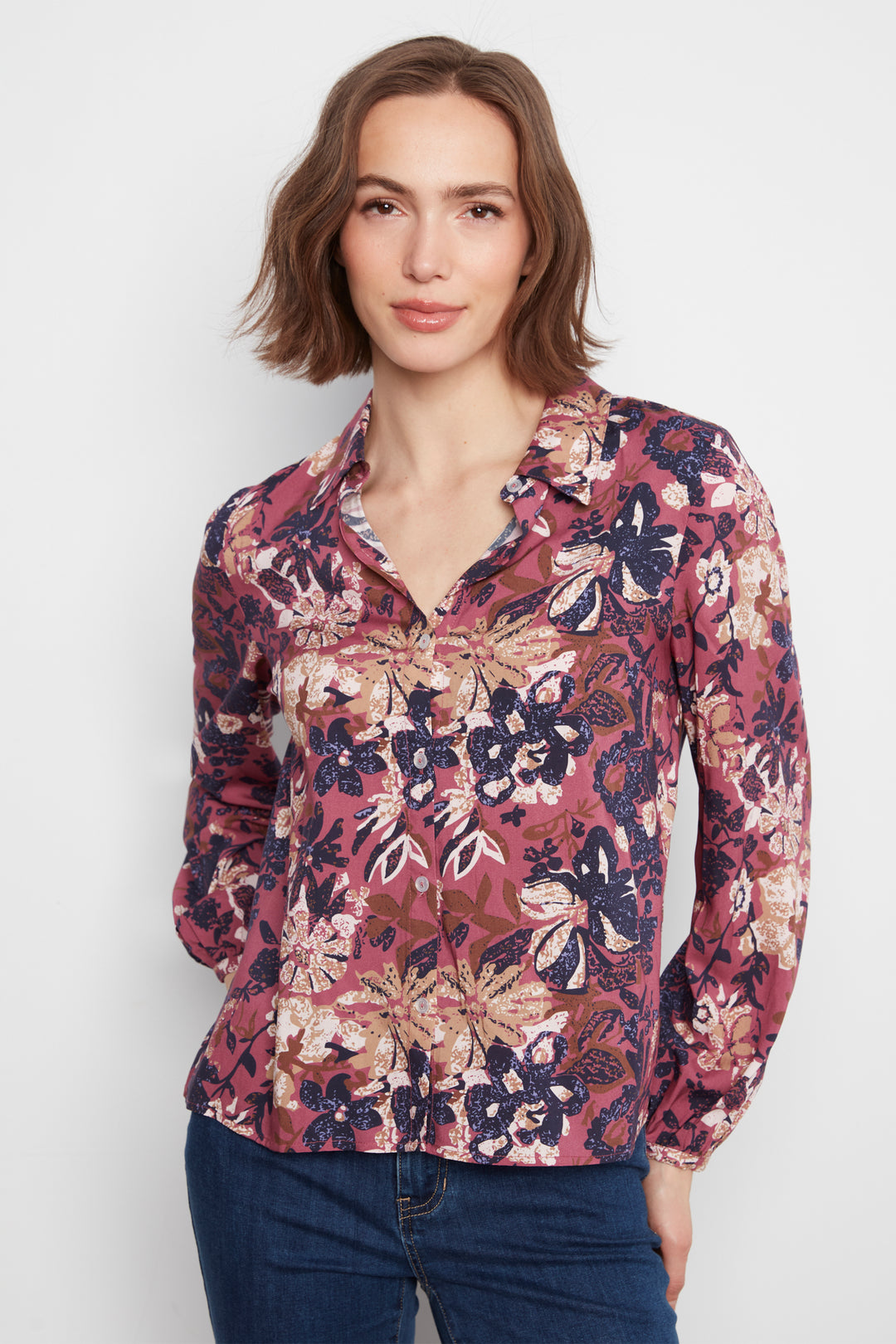 A woman wearing a long-sleeve button-up blouse with a dense floral and botanical print in dusty pink, burgundy, navy, and beige, paired with dark blue jeans, against a plain light gray background.