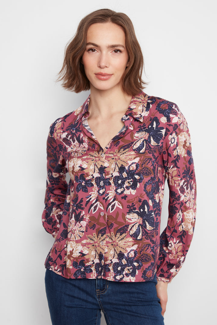 A woman wearing a long-sleeve button-up blouse with a dense floral and botanical print in dusty pink, burgundy, navy, and beige, paired with dark blue jeans, against a plain light gray background.