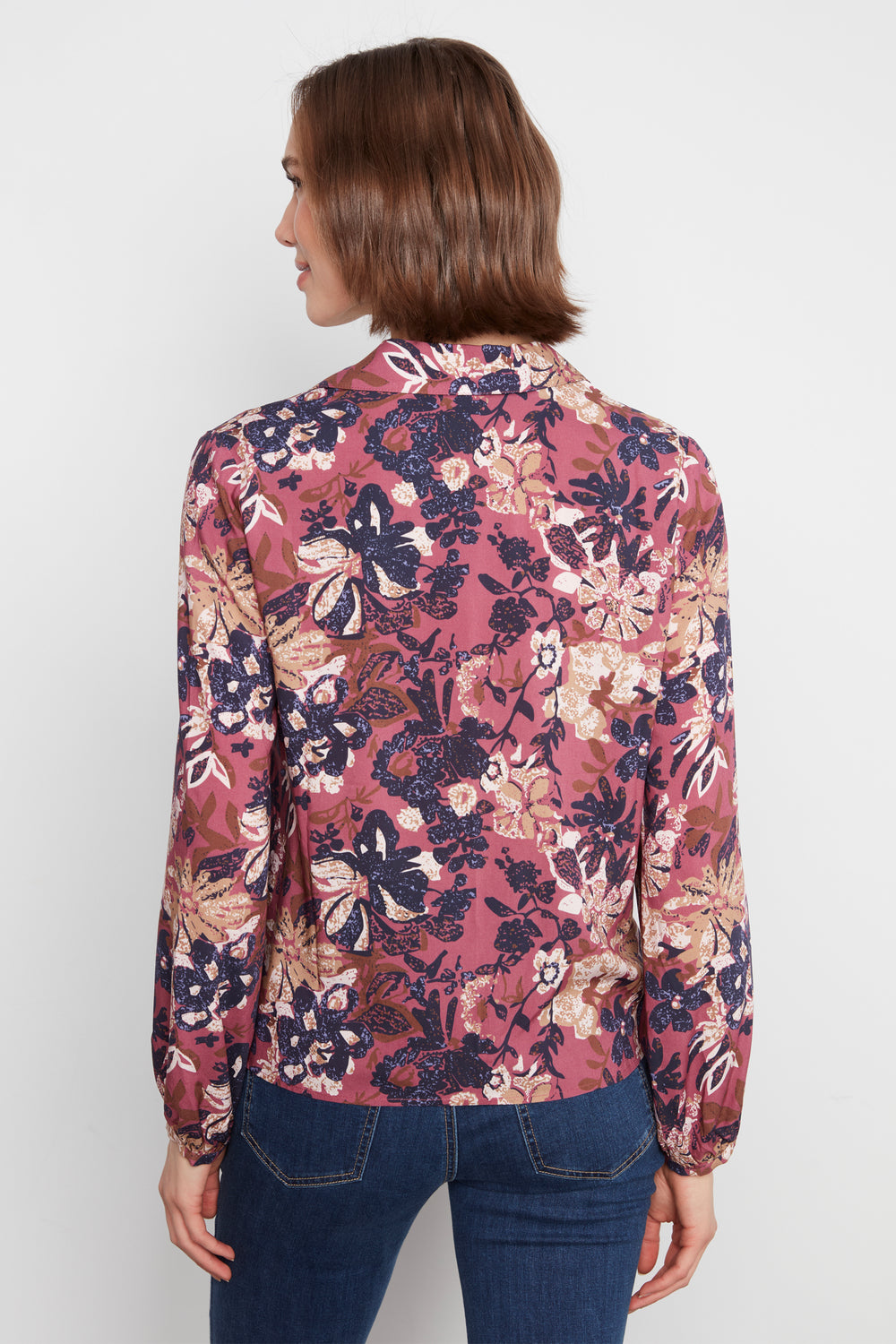 A woman shown from the back wearing a long-sleeve floral blouse with a dusty rose base and navy, beige, and brown botanical print, paired with dark blue jeans against a light gray studio background.
