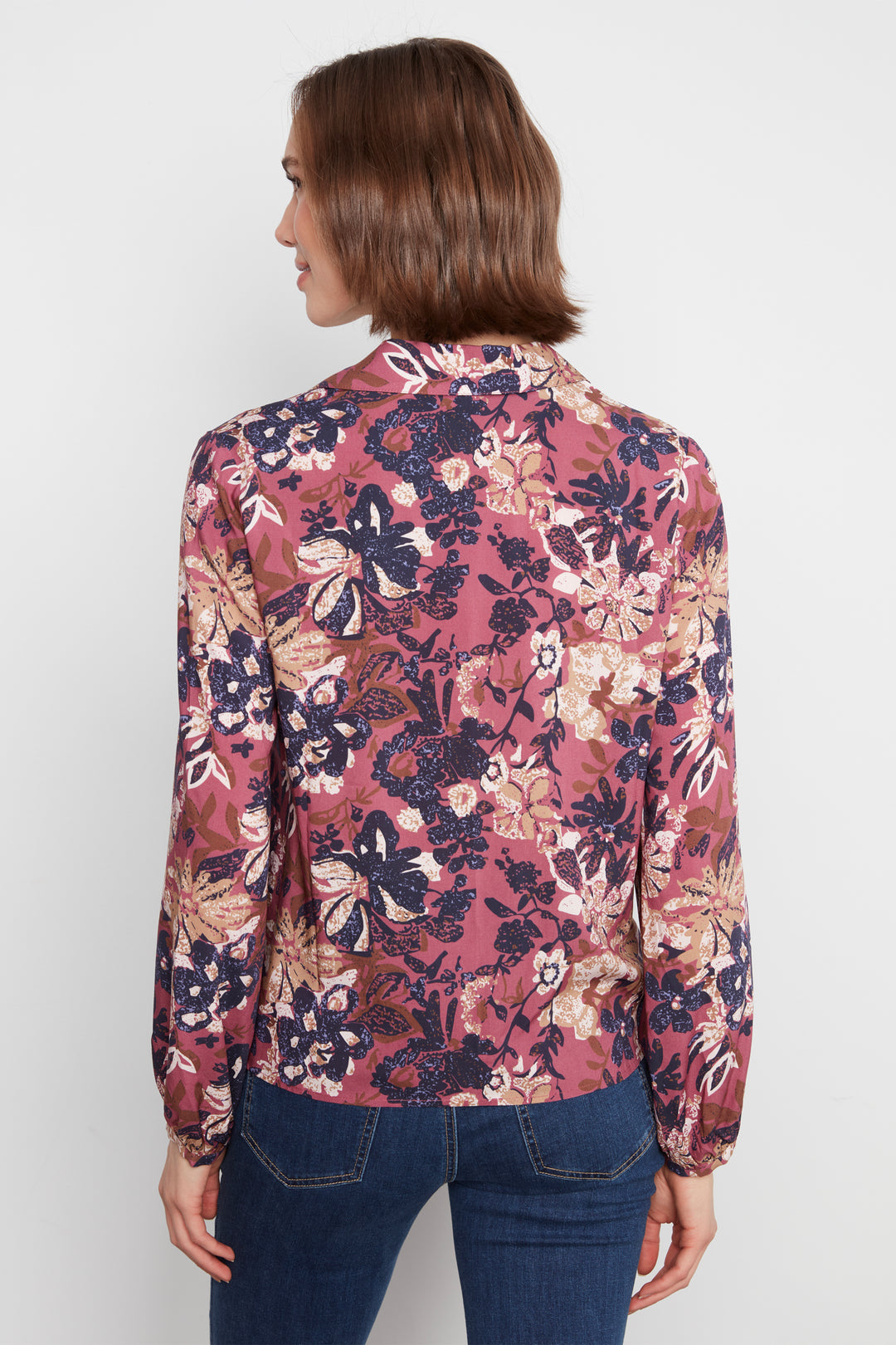 A woman shown from the back wearing a long-sleeve floral blouse with a dusty rose base and navy, beige, and brown botanical print, paired with dark blue jeans against a light gray studio background.