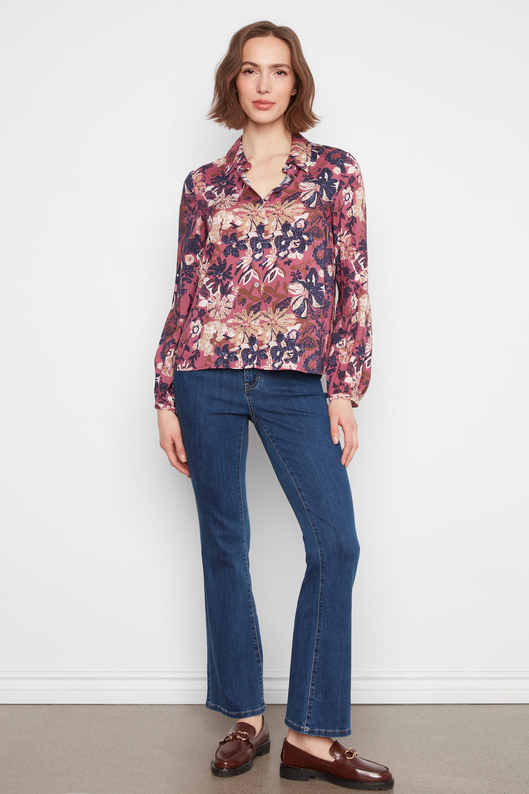 A woman modeling a long-sleeve button-up blouse with a dense floral and botanical print in shades of burgundy, pink, purple, and beige, paired with dark blue flare jeans and brown leather loafers, set against a plain white studio wall.