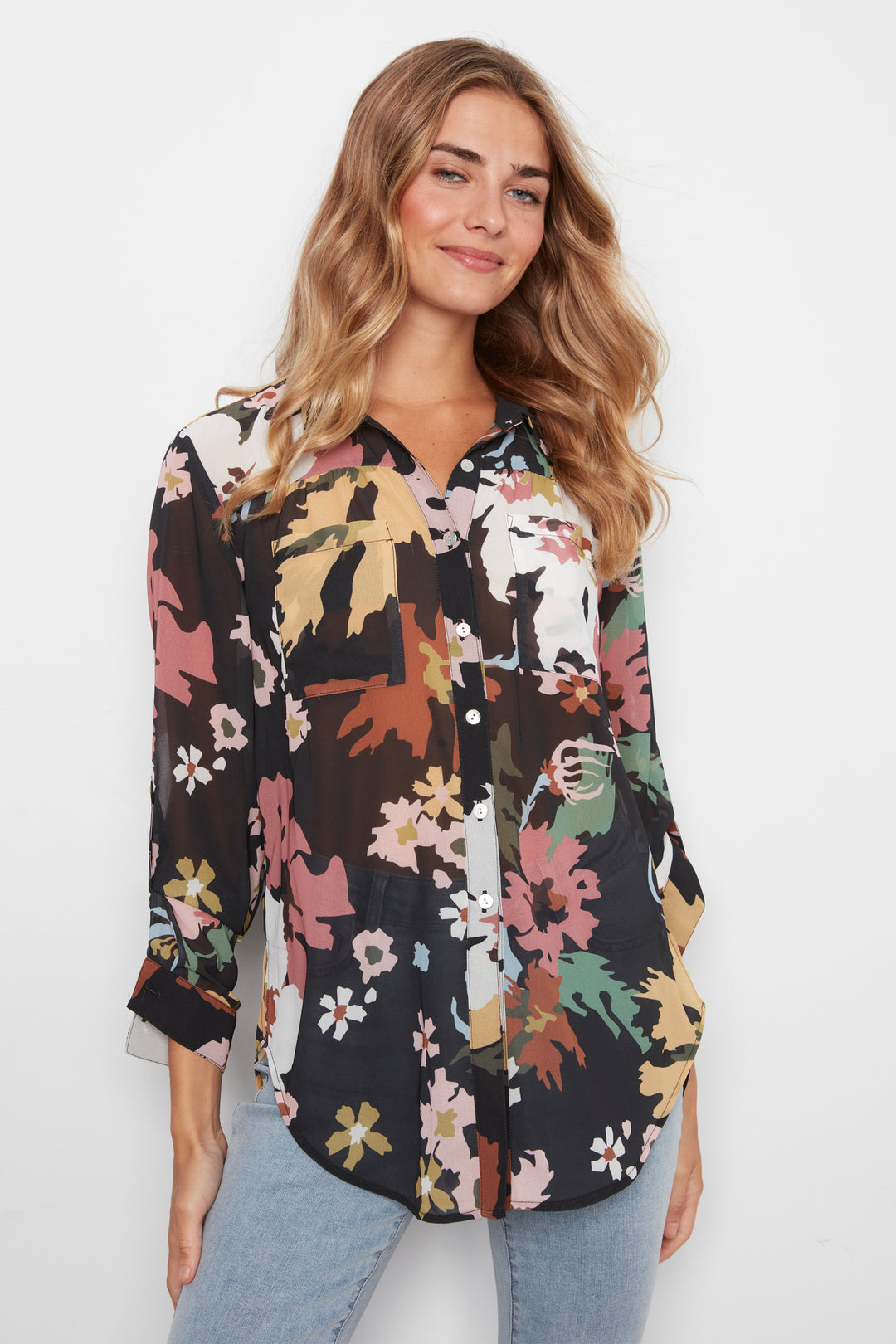 A woman wearing a black button-up blouse with a colorful floral and leaf print, paired with light blue jeans, against a plain white background.