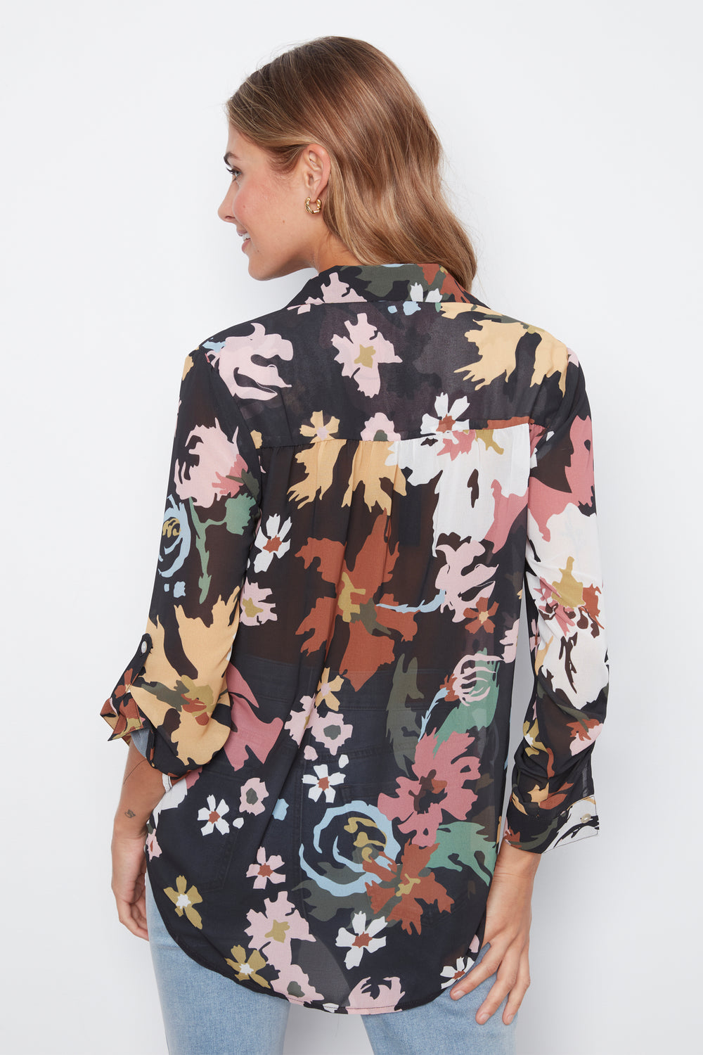 A woman shown from the back wearing a black button-up blouse with a multicolor floral and abstract print, paired with light blue jeans, against a plain white background.