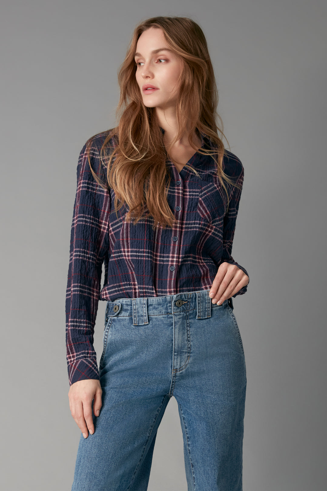 A woman modeling a navy blue long-sleeve plaid button-up shirt with red and white lines, paired with high-waisted blue jeans, against a solid gray studio background.